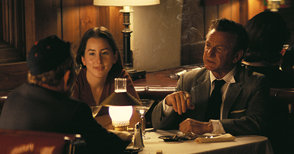 A man in a suit talks to a woman and a man in a dark cap at a candlelit restaurant table.