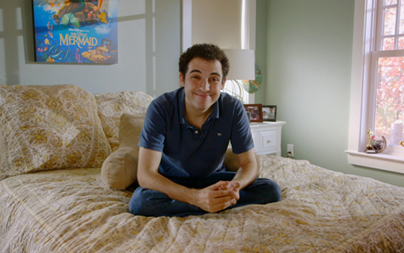 A man in a blue polo shirt sits cross-legged on a patterned bed in a bedroom.