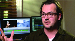 A man with glasses and dark hair gestures while speaking in front of two computer monitors showing video editing software.