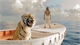 A tiger sits in a small white boat while a person stands in the background on a calm sea under a cloudy sky.