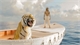 A tiger sits in the foreground of a small white boat while a person stands in the background on the water.