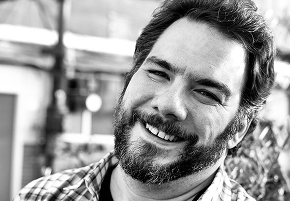 Black and white close-up portrait of a smiling man with dark hair and a beard.