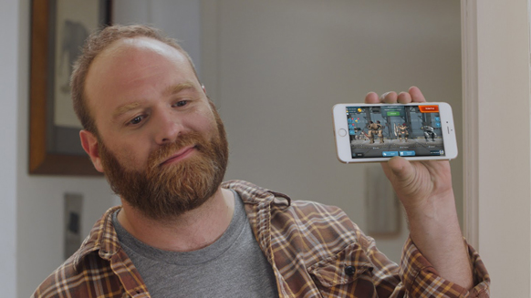 A bearded man in a plaid shirt holds up a smartphone displaying a video game.