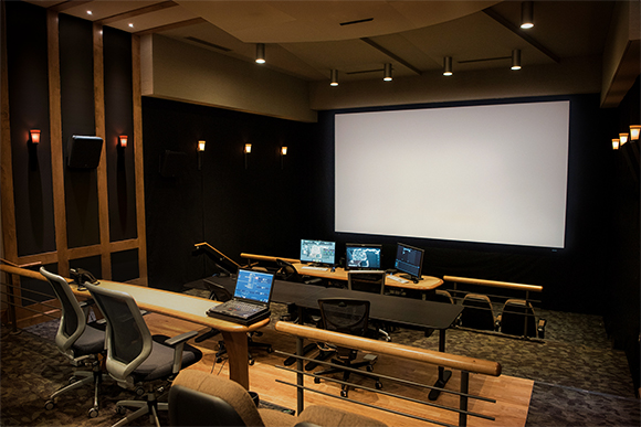 A dark control room with multiple computer monitors facing a large, blank white projection screen.