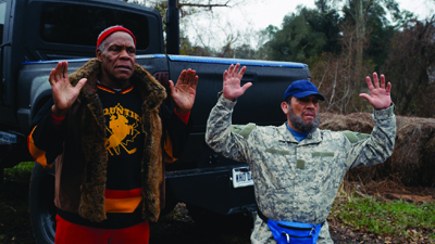Two men stand with hands raised in the air in front of a black pickup truck outdoors.