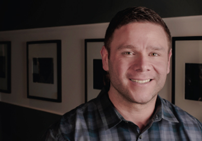A smiling man with short brown hair wearing a dark plaid button-down shirt.