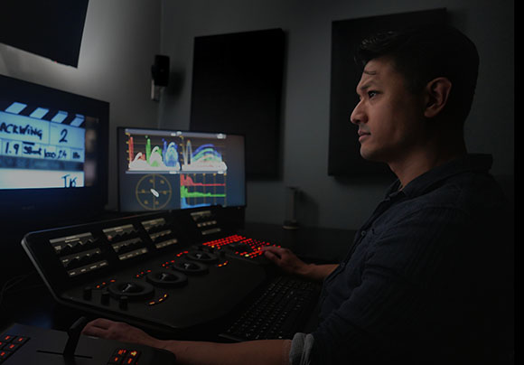 A man sits in a dark studio operating a large video editing console with multiple monitors.