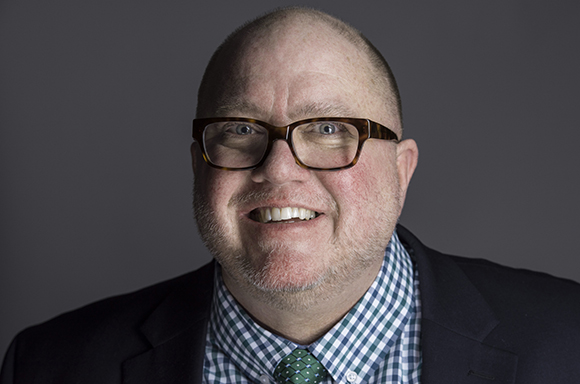 A smiling man with glasses, a beard, and a checkered shirt wearing a dark blazer.