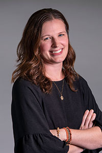 A smiling woman with wavy brown hair wearing a black top and gold necklace, posing with arms crossed.