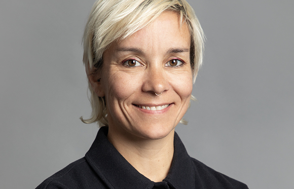 A smiling woman with short blonde hair and a nose ring looking toward the camera against a gray background.
