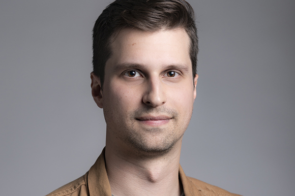 A man with short brown hair and brown eyes looking forward against a solid gray background.