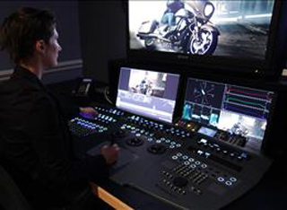 A person sits at a professional video editing desk with multiple monitors displaying footage of a motorcycle.