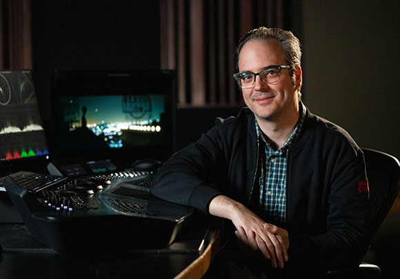 A man with glasses smiling while sitting at a dark video editing desk with monitors in a dim studio.