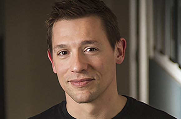 A close-up portrait of a man with short brown hair smiling against a blurred background.