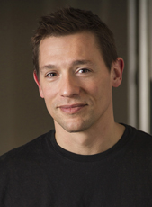 A man with short brown hair and a slight smile wearing a black crewneck shirt.