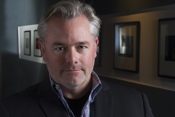 A man with salt-and-pepper hair wearing a dark blazer and a blue plaid shirt, looking directly at the camera.