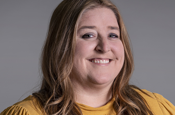 A smiling woman with long, wavy brown hair wearing a mustard yellow top against a gray background.