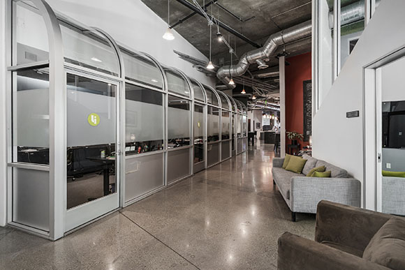 Glass-walled office cubicles line a hallway next to a grey sofa and polished concrete floor.