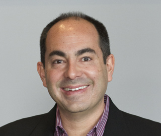 A smiling man with short dark hair wearing a black blazer and a striped collared shirt.