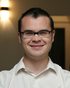 A smiling man with short dark hair and glasses wearing a white button-down shirt.