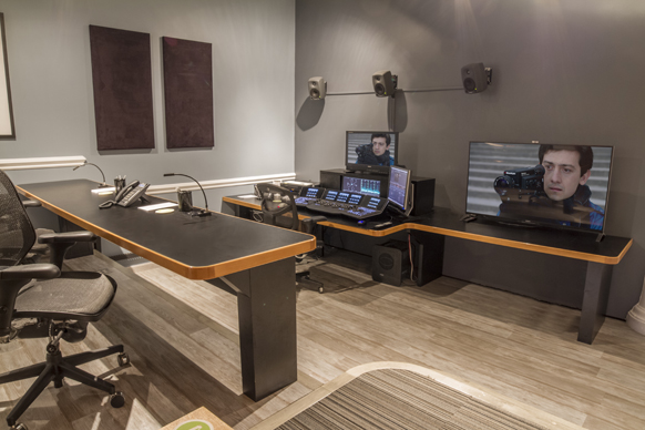 A dark-walled control room with a large desk, multiple monitors, audio speakers, and editing consoles.