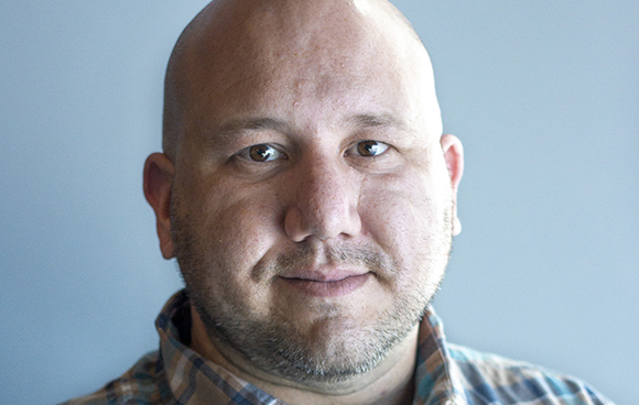 Close-up portrait of a bald man with light stubble looking directly at the camera against a plain background.
