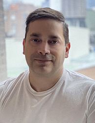 A man with short dark hair wearing a white t-shirt, looking forward against a blurred urban background.
