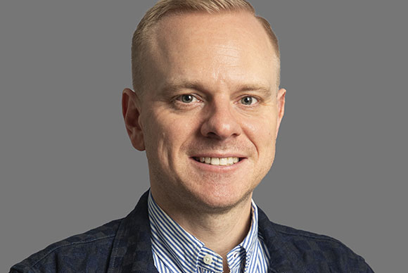 A smiling man with short blond hair wearing a blue striped shirt and a dark patterned jacket.