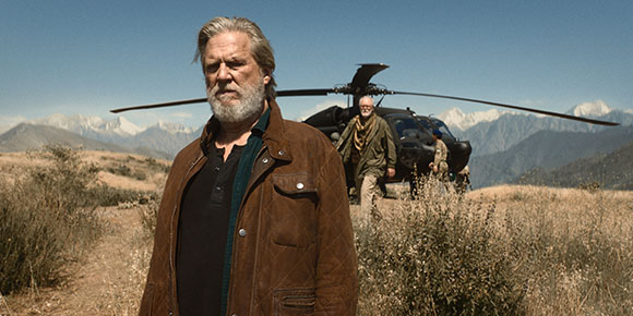 A bearded man in a brown jacket stands in a field with a black helicopter and mountains in the background.