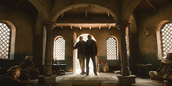Two men stand in a sunlit doorway of a stone building with arched windows and wooden pillars.