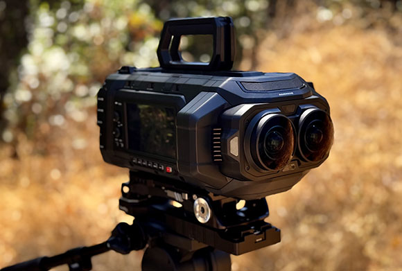 A black professional video camera with dual lenses mounted on a tripod against a blurred natural background.