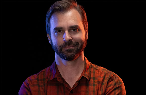 A man with dark hair and a beard wearing a red plaid shirt against a black background.