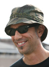 A smiling man wearing a camouflage bucket hat and dark sunglasses.
