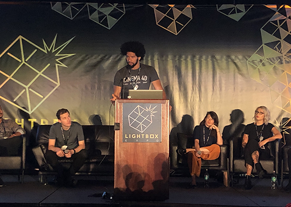 A man with an afro speaks at a podium with the Lightbox Expo logo. Behind him, four people are seated on a couch.