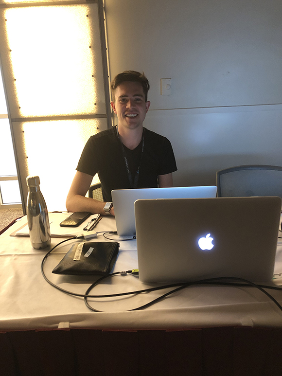 A man wearing a black t-shirt and a lanyard smiles at the camera while sitting behind a table with two open laptops, a metal