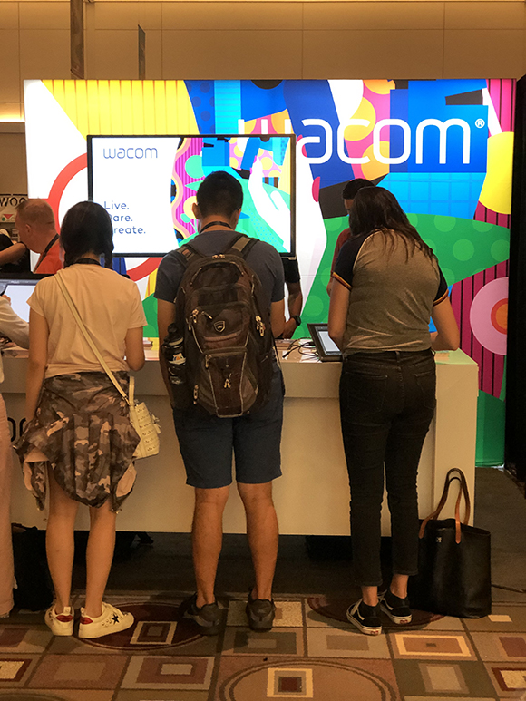 Three people stand at a Wacom booth, looking at displays and interacting with products. A large Wacom logo is visible on the