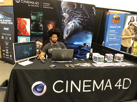 A man with an afro sits at a table with Cinema 4D branding, working on a laptop and computer. The table displays promotional