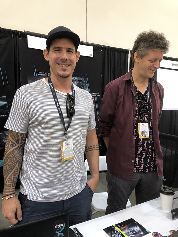 Two men stand at a table with lanyards around their necks. The man on the left smiles at the camera, wearing a striped shirt