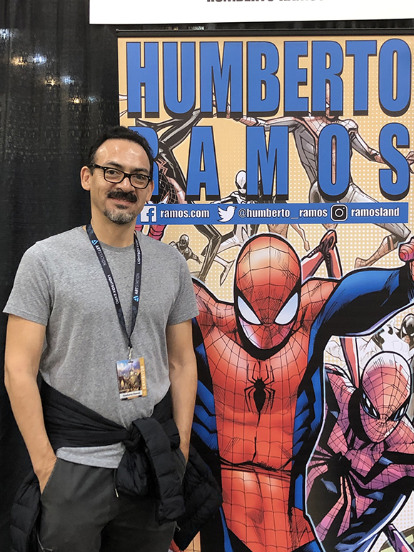 A man with glasses and a mustache stands in front of a banner with the name Humberto Ramos and illustrations of Spider-Man.