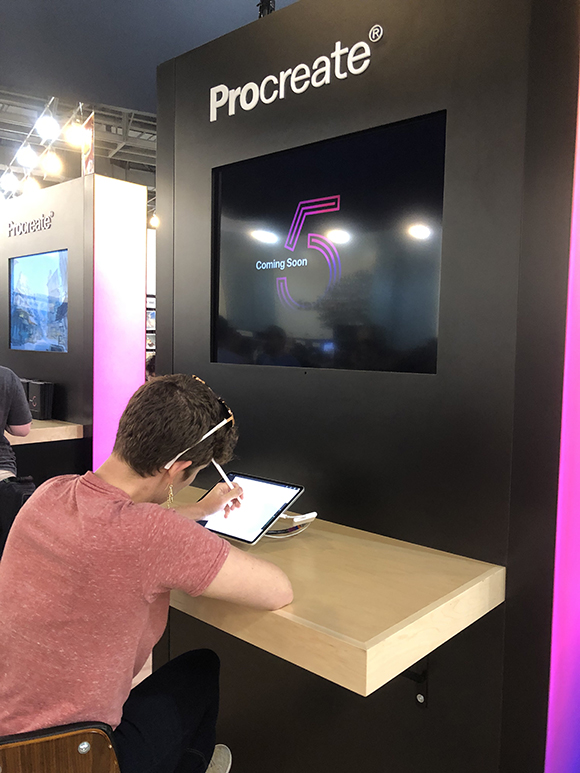 A person draws on a tablet at a Procreate booth. A large screen behind them shows "Procreate 5 Coming Soon" with a stylized n