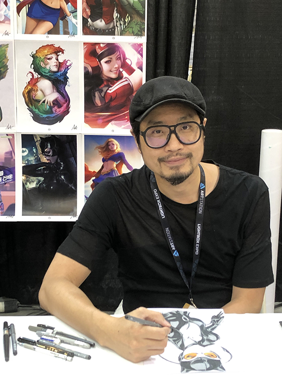 An artist wearing glasses and a cap draws on a piece of paper at a table. His artwork is displayed on the wall behind him.