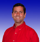 A smiling man with dark hair wearing a red button-down shirt against a blue gradient background.