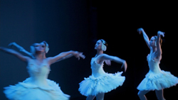 Three ballet dancers in white tutus performing a dance on a dark stage.
