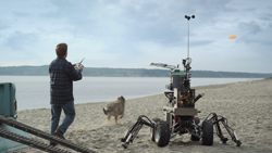 A man stands on a beach holding a remote, facing a multi-legged robot and a dog near the water.