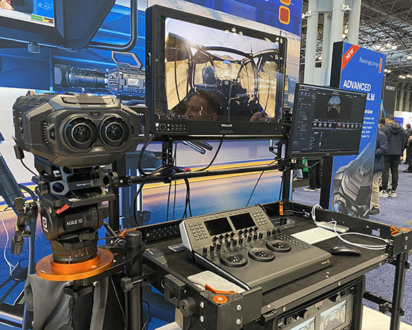 A professional cinema camera on a tripod next to a workstation with multiple monitors and control panels.