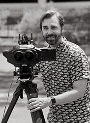 A smiling man in a patterned shirt stands behind a large professional camera mounted on a tripod.