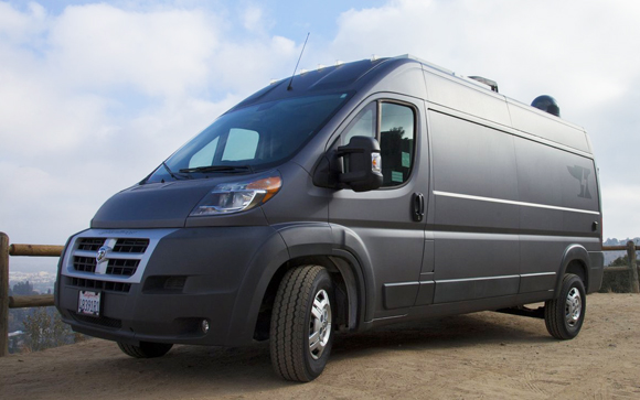 A matte black Ram ProMaster van parked on a dirt path under a cloudy sky.