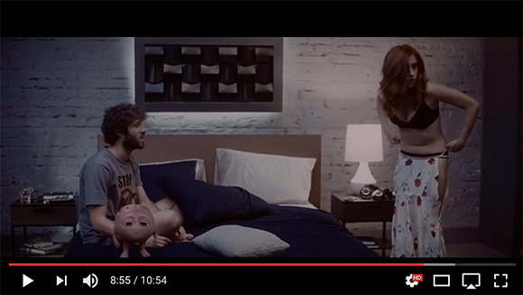 A man sits on a bed next to a small doll while a woman stands nearby in a dimly lit bedroom.