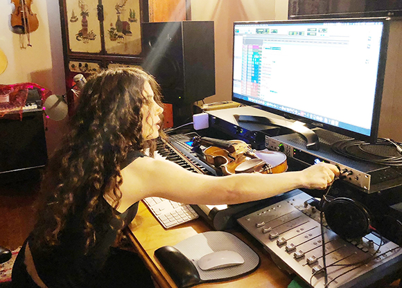 A woman with long dark hair adjusts a mixing console in front of a computer monitor and musical instruments.