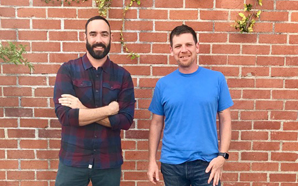 Two men smiling while standing in front of a red brick wall with climbing vines.